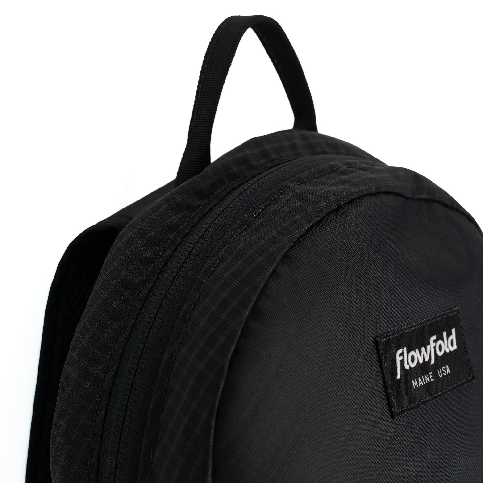 Close-up of a black Flowfold backpack showing the top handle, zipper, and a black brand patch with white text on a white background.