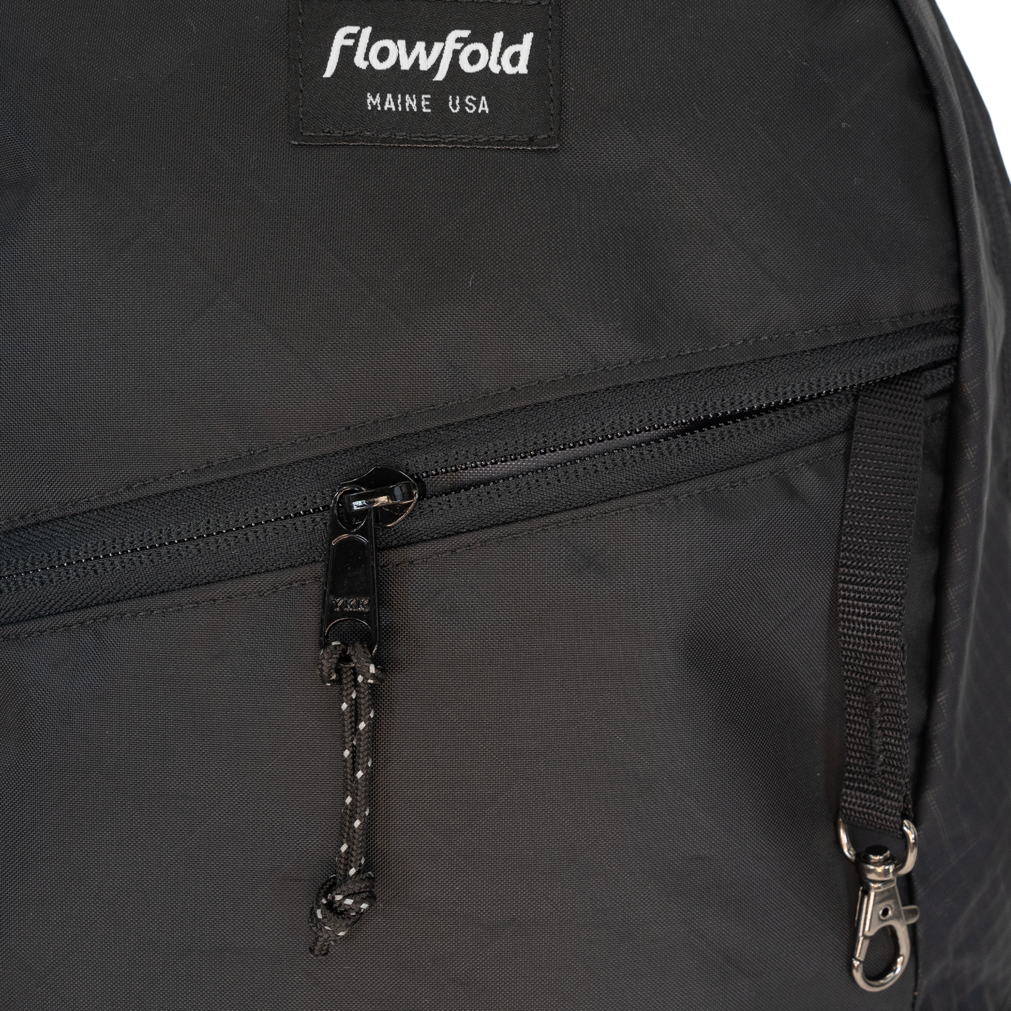 Close-up of a black Flowfold backpack showing a front zippered pocket with a black zipper pull, a silver-tone carabiner, and the Flowfold brand patch on the upper left.