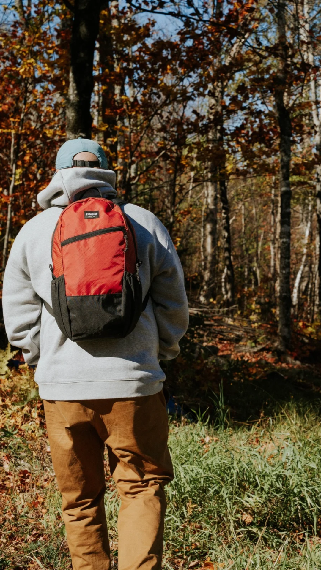 A person viewed from behind wearing a red and black backpack, a light gray hoodie, and brown pants while walking through an autumn forest.