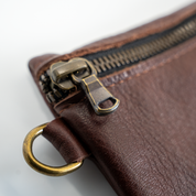 Close-up of a brown leather pouch with a brass zipper and ring on a white background