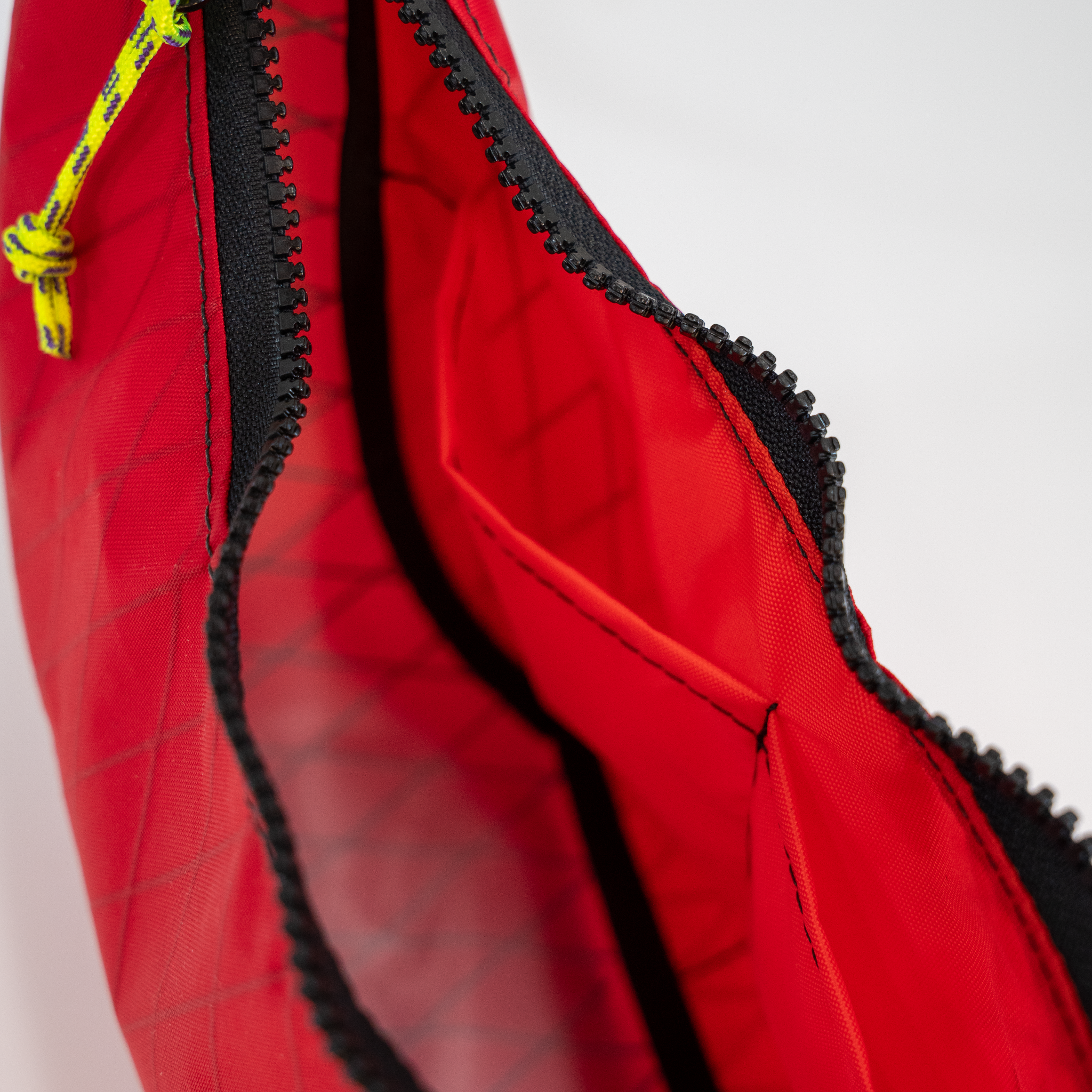 Close-up of a red bag with black zipper trim and a small yellow-and-black cord detail, shown against a light gray background.