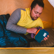 Man in a tent holding a blue and purple fleece grid belt bag with black strap, stuffing it with a sambob hoodie