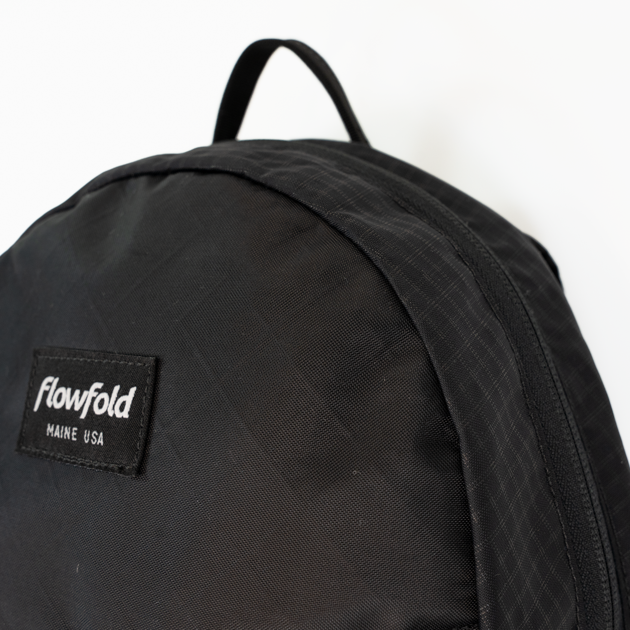 Close-up of a black Flowfold backpack showing the top handle, textured fabric, and a black brand patch with white text reading 'Flowfold MAINE USA' on a white background.