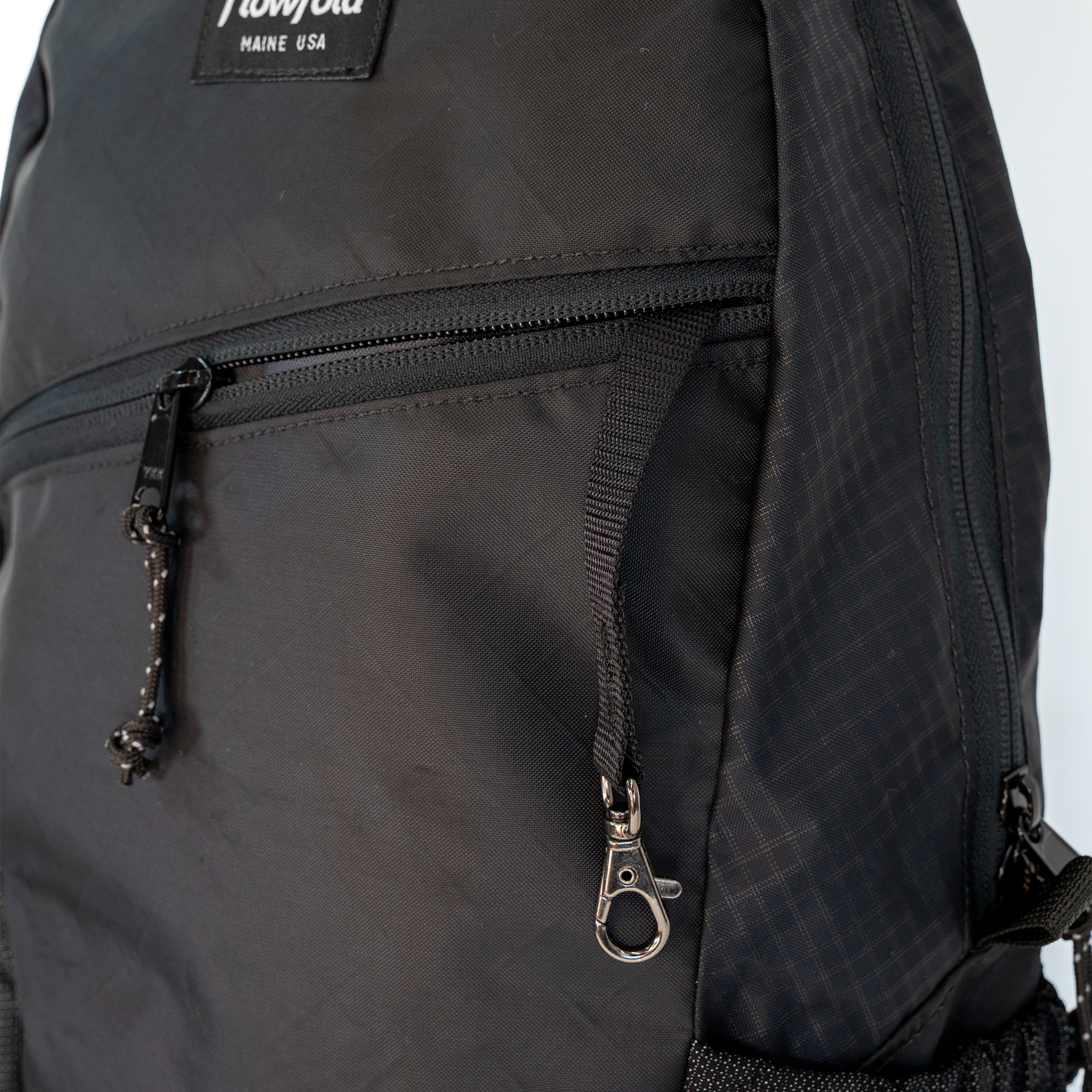 Close-up of a black backpack showing a front zippered pocket with a silver-tone lobster clasp and a black brand patch reading 'Flowfold MAINE USA' at the top.