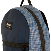 Close-up of a Flowfold backpack showing a navy blue front panel, a blue-and-white grid patterned accents, black zippers, and a black Flowfold brand patch.